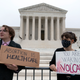Two protestors stand outside the Supreme Court building holding signs. One says "abortion is healthcare," the other says "you are waking a volcano."